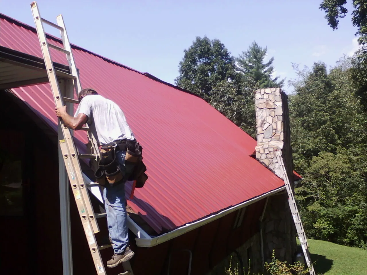 Roofer on a ladder beside a red metal roof and stone chimney — Soffit & Fascia Repair by Roofing Brothers in Sauk City